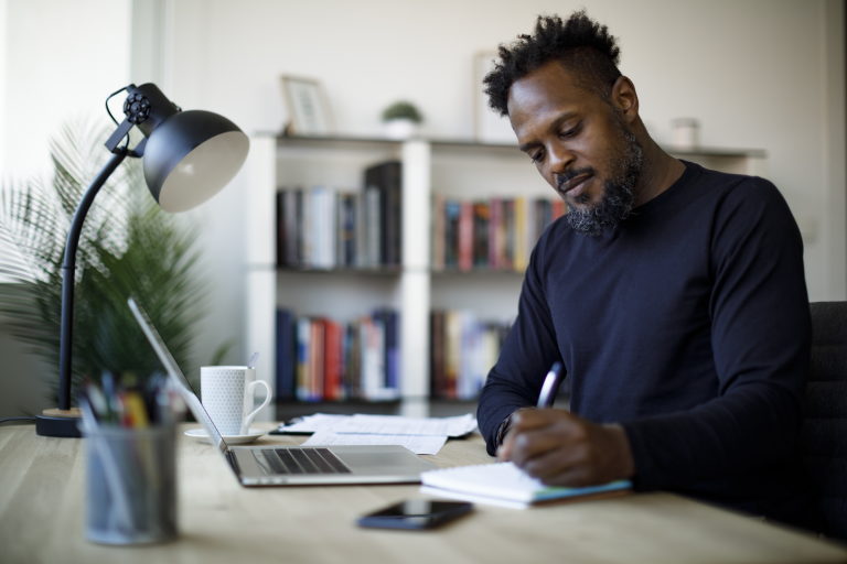 Man achter laptop schrijft in kladblok.